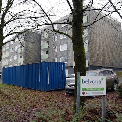Blick auf ein Mehrfamilienhaus, davor ein Parkplatz und ein großer Müllcontainer.