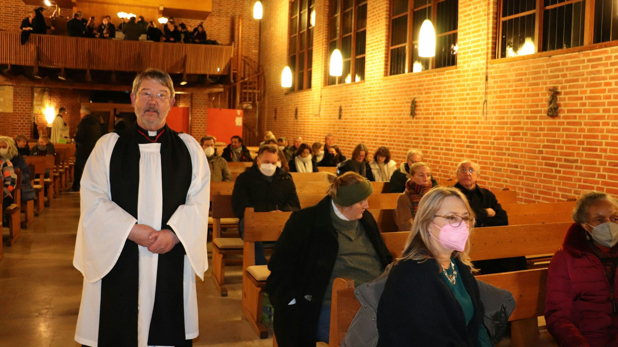 Ein grauhaariger Mann mit Schnauzer steht im weißen Messgewand und einem schwarzen Schal in einer Kirche inmitten von halb besetzten Kirchenbänken.