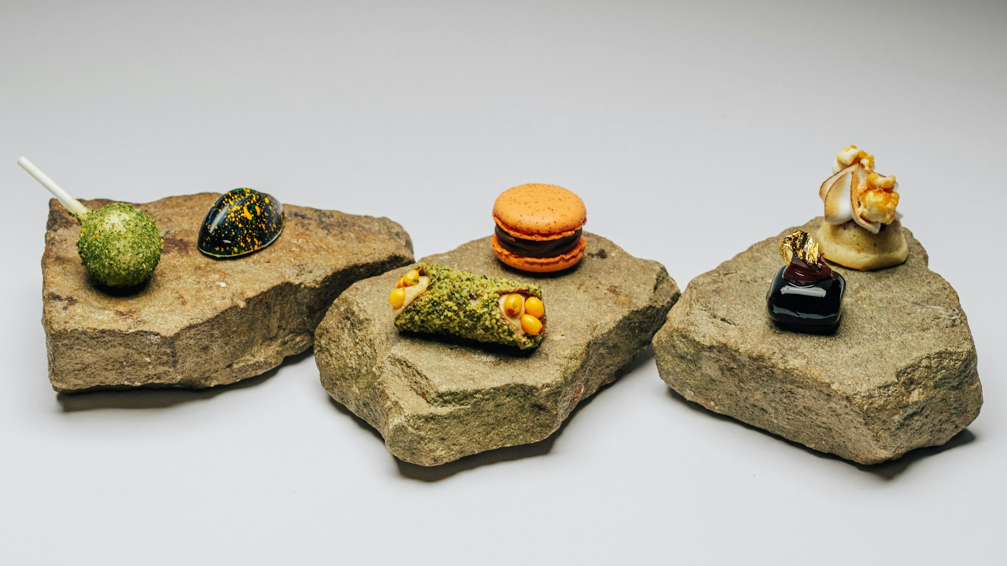 Sechs kleine Desserts werden im Ox & Klee in Köln auf drei Steinplatten serviert. Dabei ist unter anderem eine Art grüner Lolli, eine schwarze Praline mit orangenen Sprenkeln oder ein orangene Macaron.