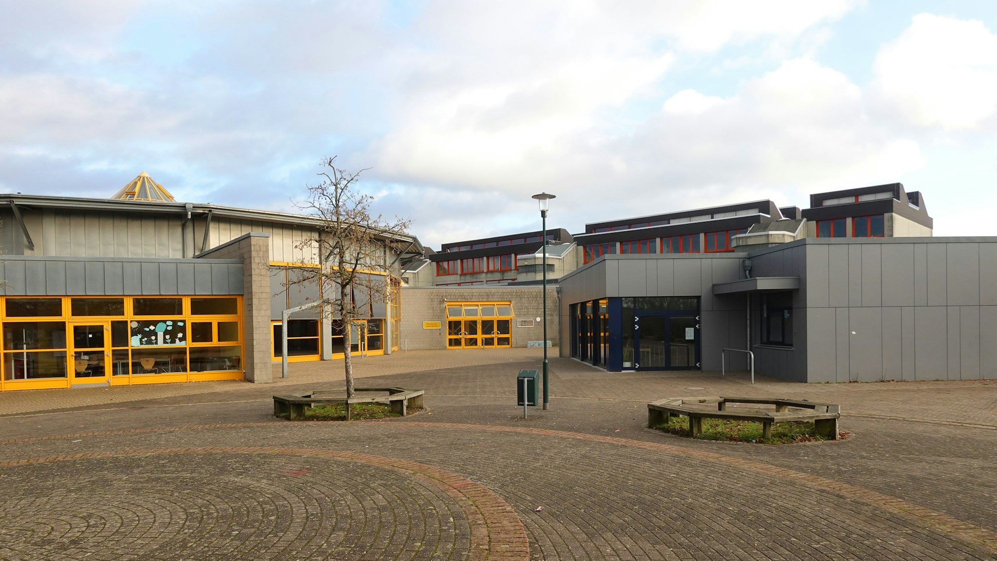 Eine graue Gesamtschule mit orangefarbenen Fensterläden. Vor der Schule stehen zwei Bänke