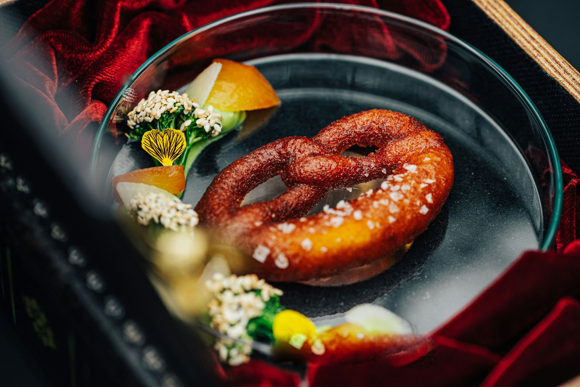 Eine Bretzel liegt im Ox & Klee auf einer Glasplattte, fein angerichtet daneben liegen ein paar Kräuter und Blüten.