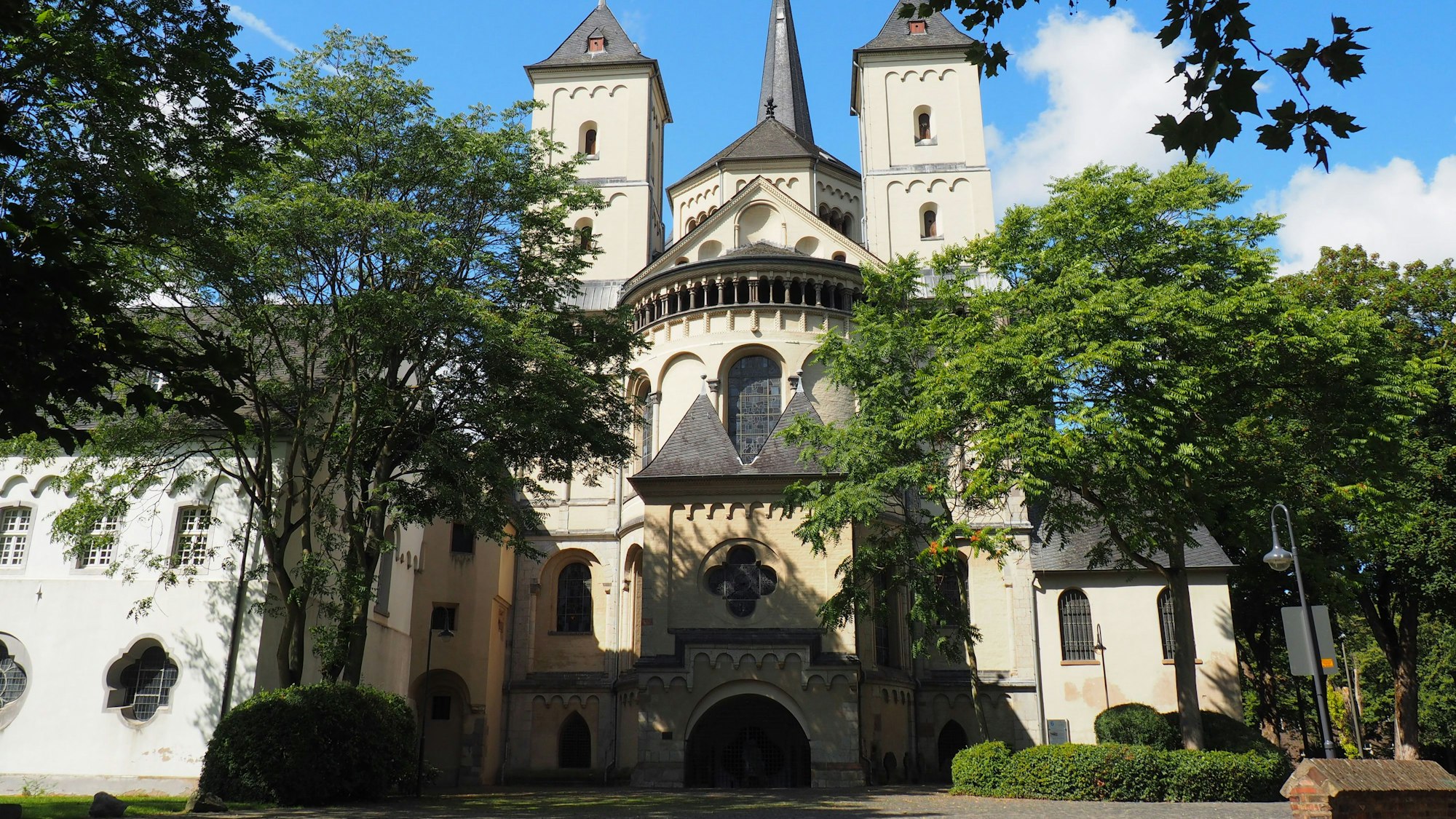 Im Hintergrund sieht man eine Abtei mit den grauen Dächern. Links und rechts wachsen Bäume.