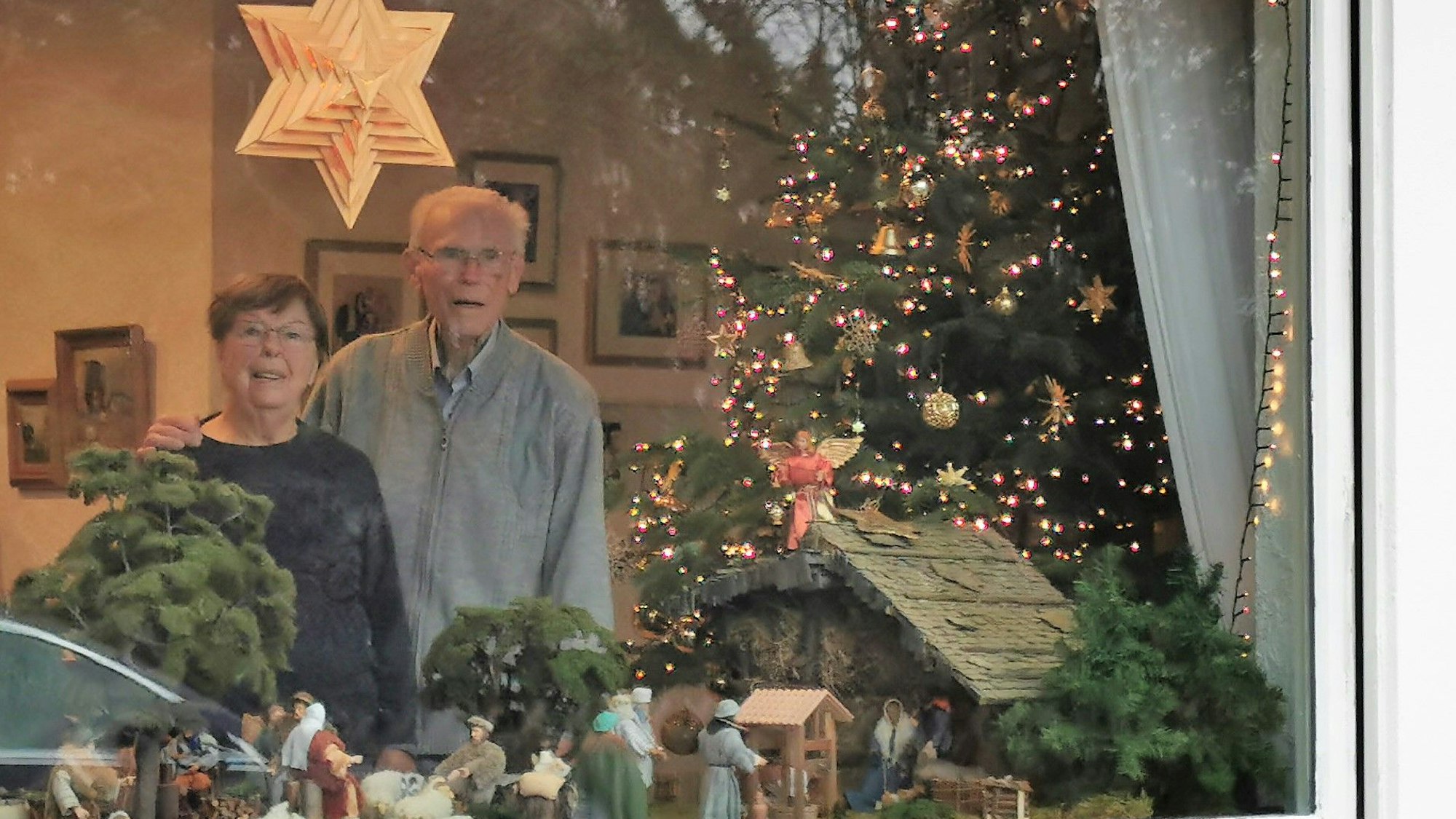 Ein älteres Paar steht hinter dem Fenster. Im Schaufenster sieht man eine handgemachte Krippe.