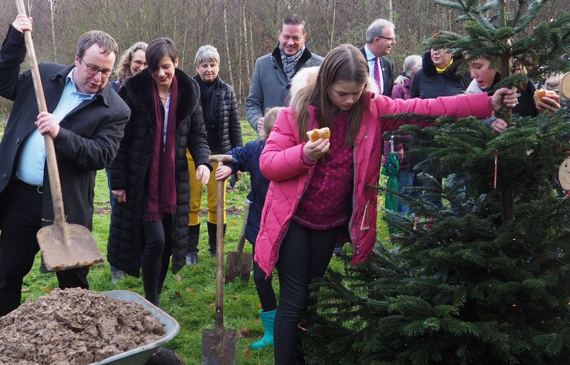 Viele Menschen stehen um einen Tannenbaum herum. Ein Mann schaufelt Erde aus einer Schubkarre.