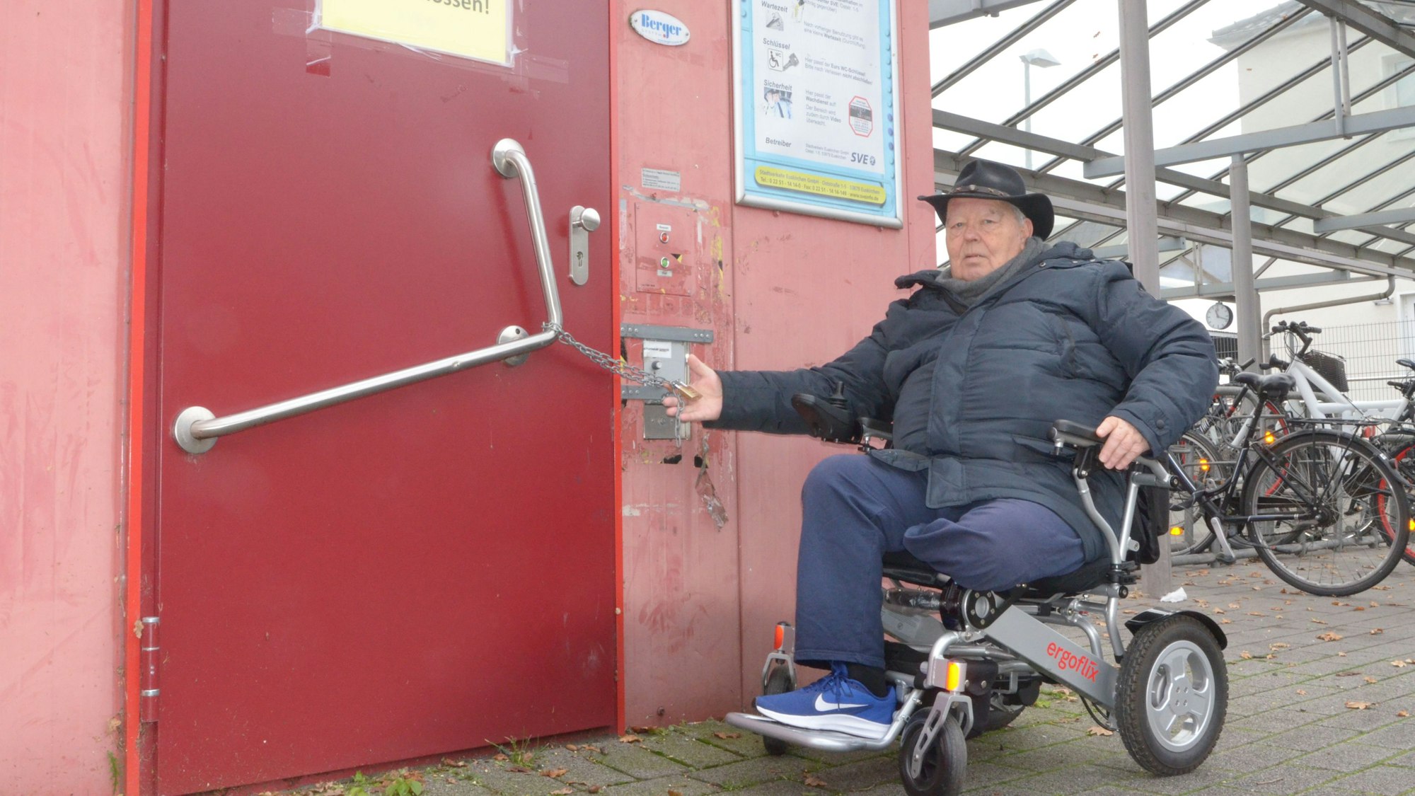 Der Rollstuhlfahrer Wilhelm Hachenberg vor einer geschlossenen Toilette am Bahnhof Euskirchen. An der Tür hängt eine Kette mit einem Vorhängeschloss.