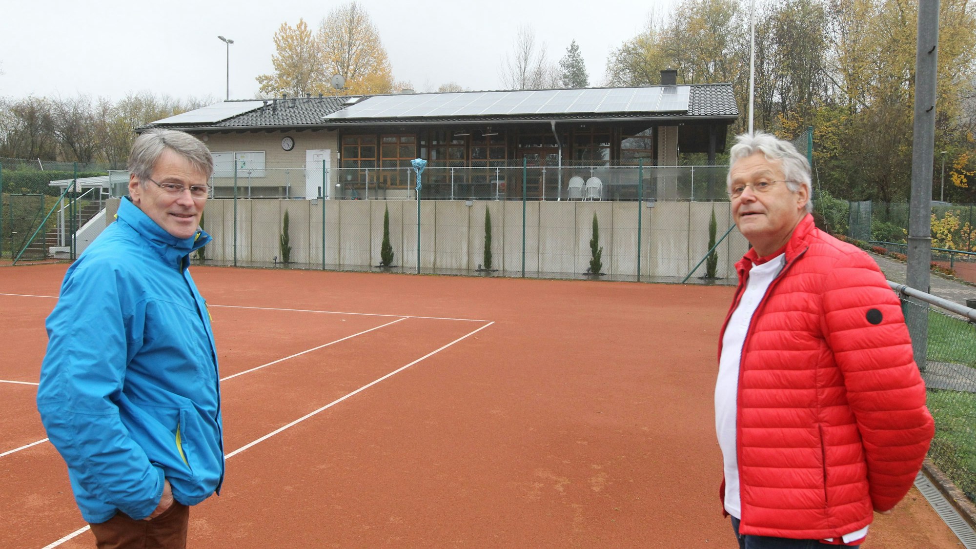 Zwei Männer in blauer und roter Jacke stehen auf einem Tennisfeld.