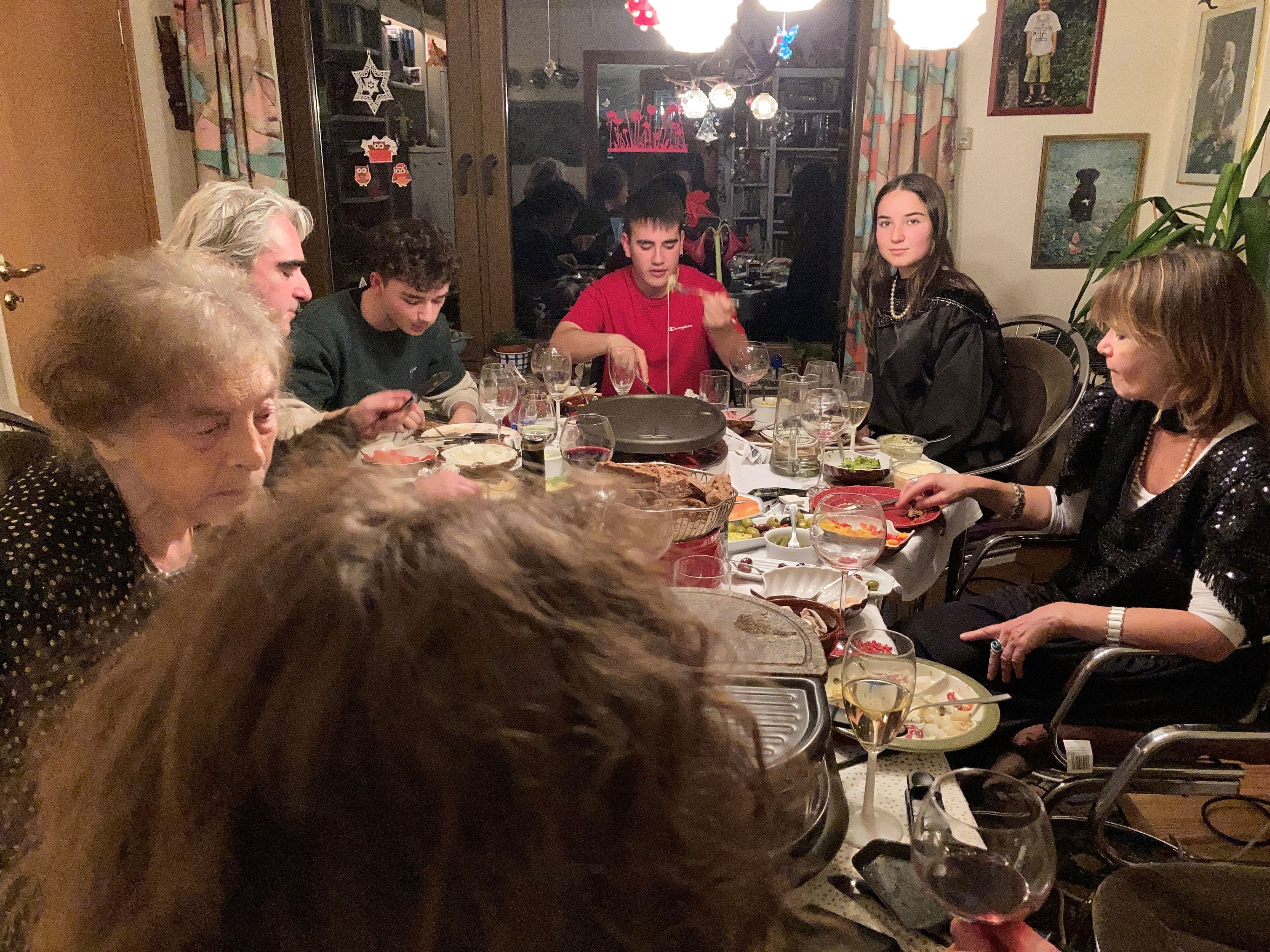 Sieben Menschen sitzen einem reich gedeckten Tisch mit einer Raclette-Platte.