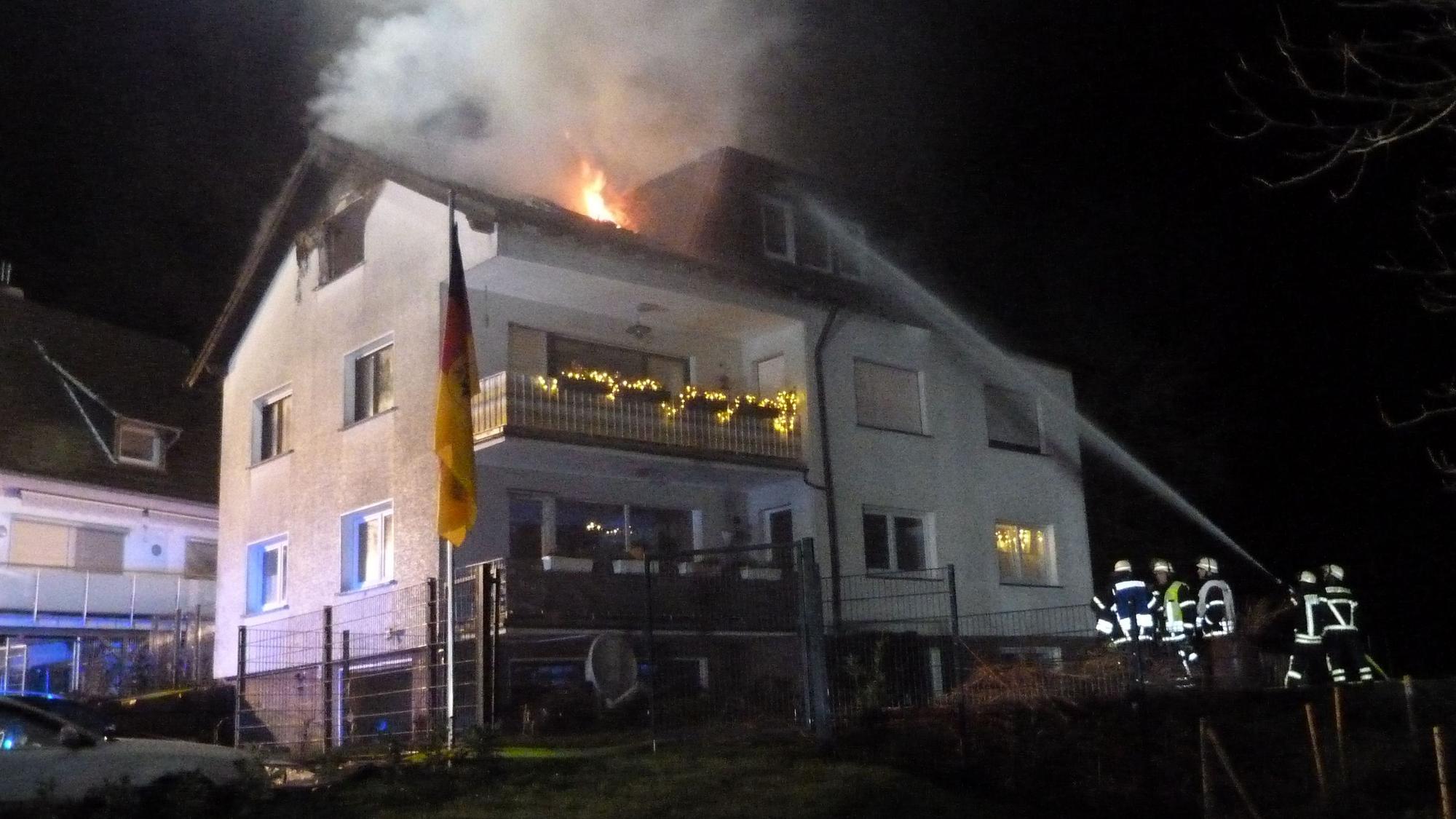 Aus dem Dach eines Hauses schlagen Flammen. Feuerwehrleute halten mit einem Löschwasser-Schlauch darauf.