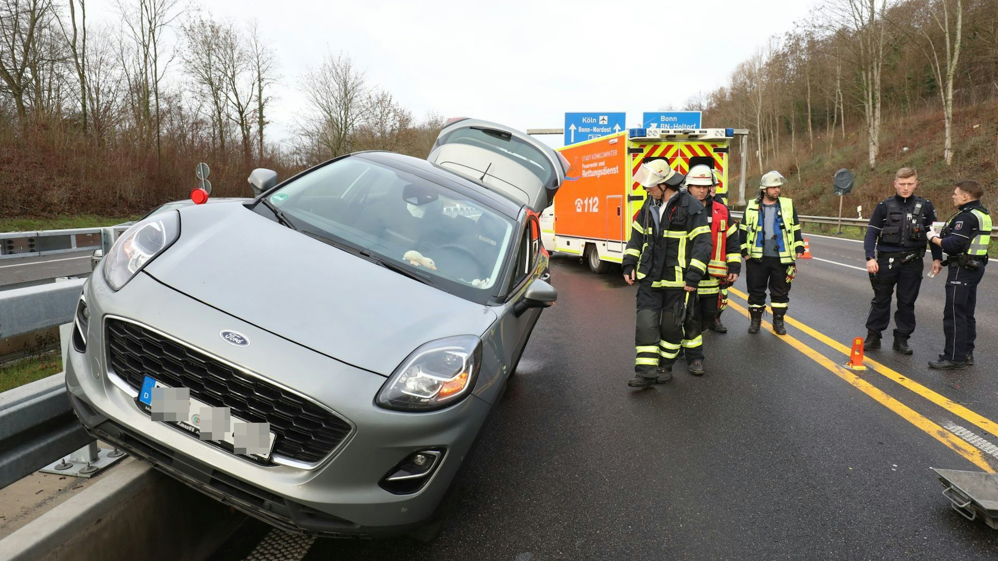 Ein silbernes Auto hängt halb auf einem Betonsockel mit der Leitplanke und halb auf der Fahrbahn. Feuerwehrleute und Polizisten stehen an der Unfallstelle.