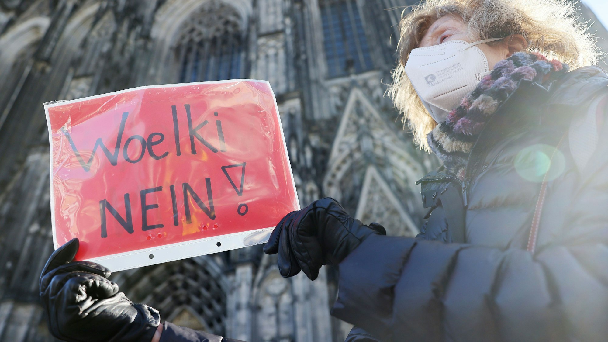 Eine Frau nimmt mit einem Plakat mit der Aufschrift „Woelki Nein!“ an einer Kundgebung der kirchenkritischen Bewegung Maria 2.0 gegen Kardinal Rainer Maria Woelki teil. Im Hintergrund ist der Kölner Dom zu sehen.
