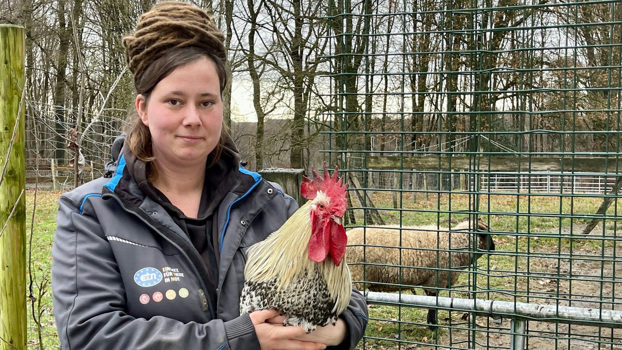 Eine Frau mit einem großen Dutt hält einen Hahn im Arm. Sie trägt einen Arbeitsoverall, im Hintergrund steht ein Schaf hinter einem Zaun.