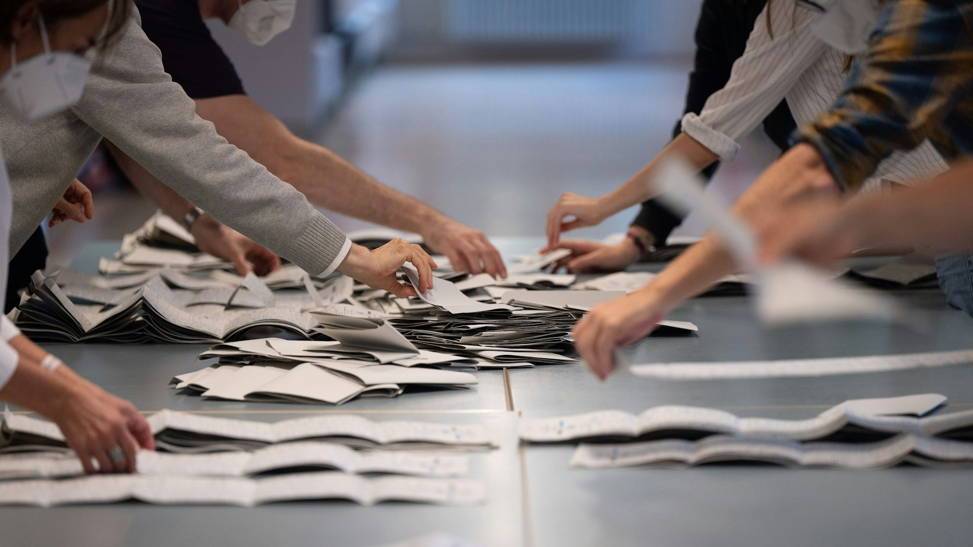 Hände greifen von rechts und links nach Wahlzetteln auf einem Tisch.