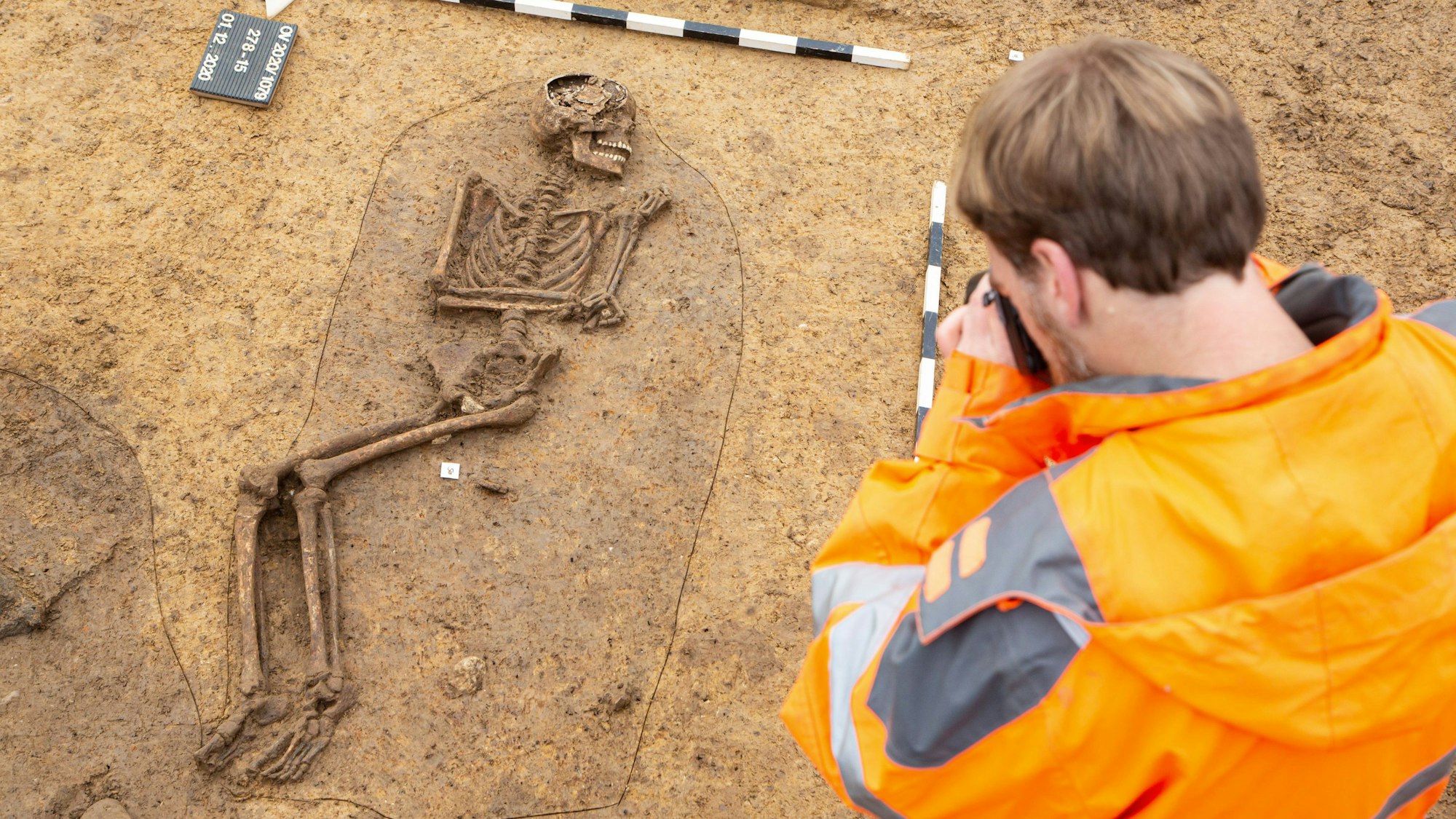 Oedekoven, archäologische Untersuchungen auf der Baustelle der christlichen Schule von der Firma Archaeonet. Hier: Fund eines spätrömischen Skeletts, das sehr gut erhalten ist (eines von vieren auf dieser Fläche, das am besten erhalten ist). Vermutlich junge Person, Geschlecht vor der Untersuchung eines Anthropologen noch unklar, wurde in einer kleinen Grube bestattet, deshalb die Anwinklung der Beine. Unklar ist noch, warum die Arme so angewinkelt sind; bei dem daneben gefundenen Skelett war das auch so. Normalerweise befinden sich die Hände vor dem Becken. Direkt daneben befindet sich eine dunkle Erdschicht, die auf einen großen Hauspfosten einer mittelalterlichen Grube hinweist. Hier fotografiert Grabungsleiter Felix Kunze das Skelett um möglicherweise ein 3D-Modell davon zu machen.