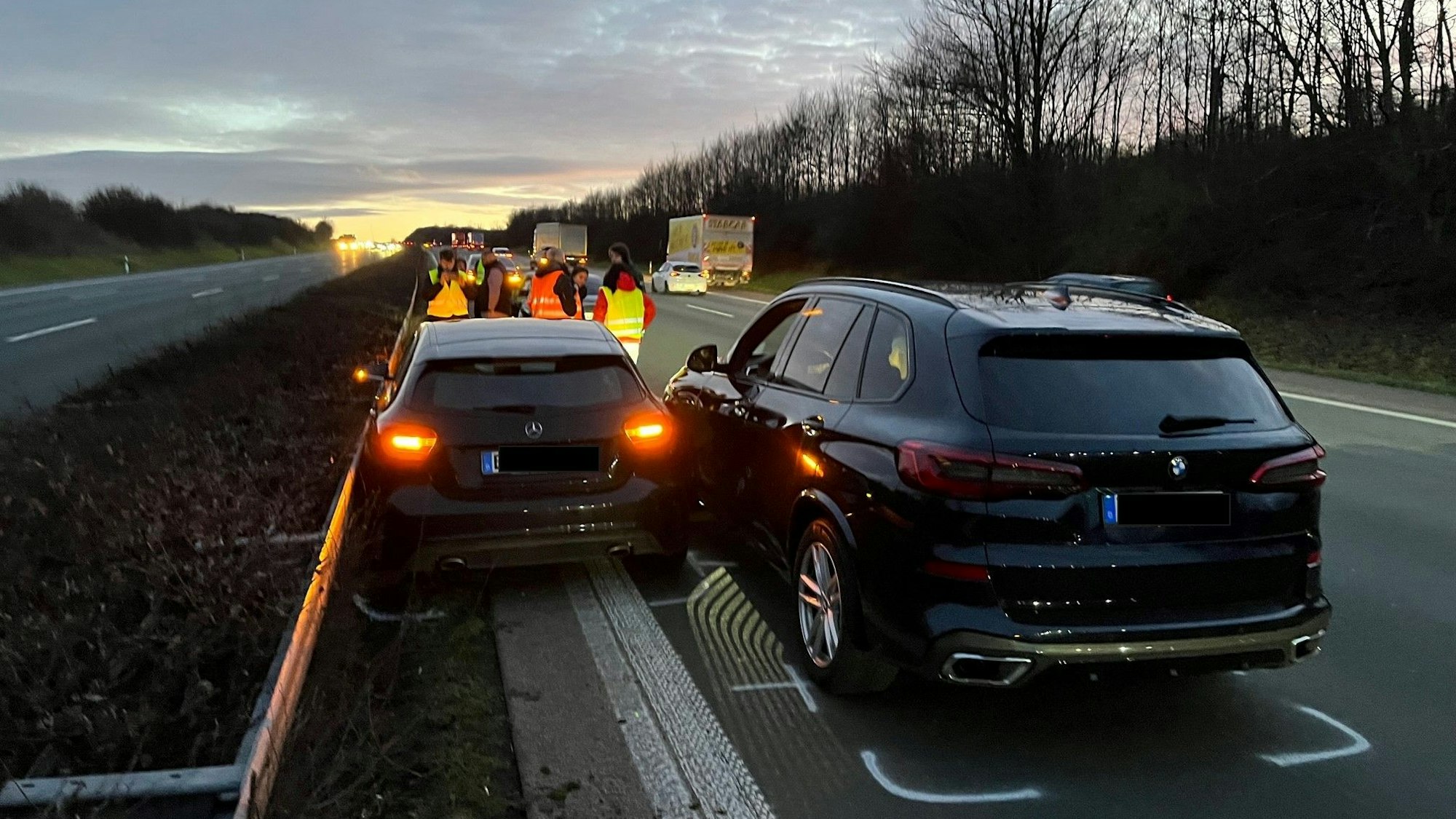 Zwei dunkle Fahrzeuge, ein Mercedes und ein BMW, stehen nach einem Zusammenstoß Blech an Blech auf der Autobahn neben der Mittelleitplanke. Menschen in Warnwesten stehen dahinter.