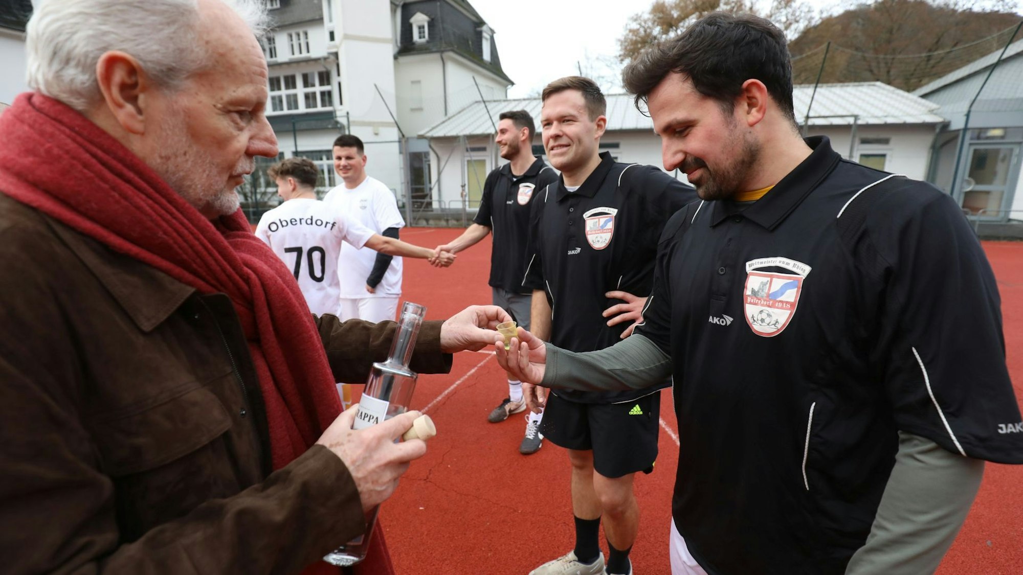 Ein Mann mit rotem Schal und brauner Jacke (Schiedsrichter Bernd Baumgarten) reicht einem Fußballspieler in schwarzem Trikot auf dem Kleinspielfeld am Haus Rheinfrieden ein Gläschen mit Schnaps.