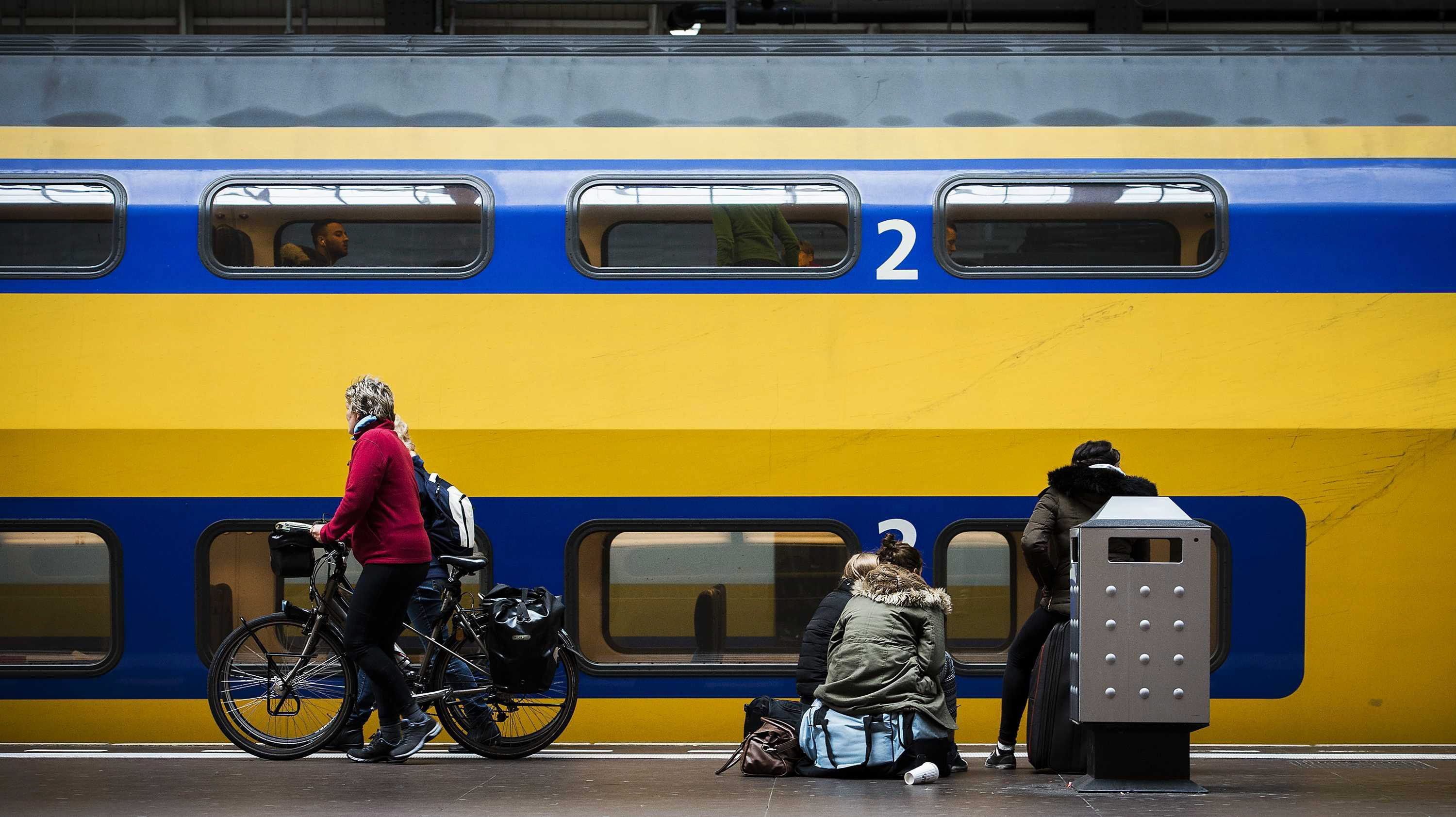 Ein Zug hält an einem Bahnhof in den Niederlanden. Vor dem Zug warte Passagiere, eine Frau in einer roten Jacke schiebt ein Fahrrad am Gleis entlang. (Archivbild)