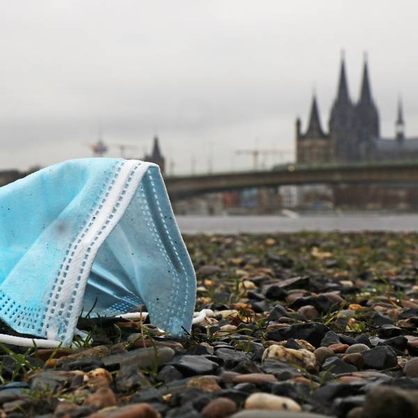 Eine weggeworfene Maske liegt am Rheinufer auf dem Boden. Im Hintergrund sieht man unscharf den Dom.