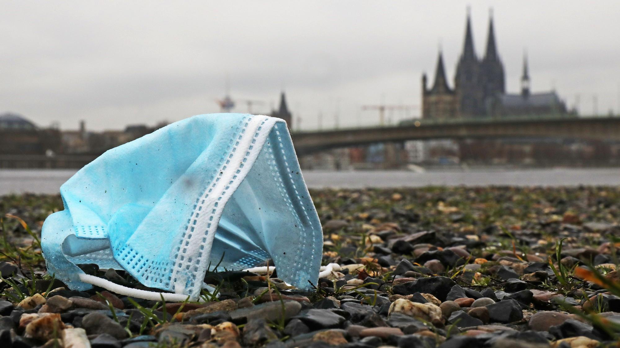 Eine weggeworfene Maske liegt am Rheinufer auf dem Boden. Im Hintergrund sieht man unscharf den Dom.