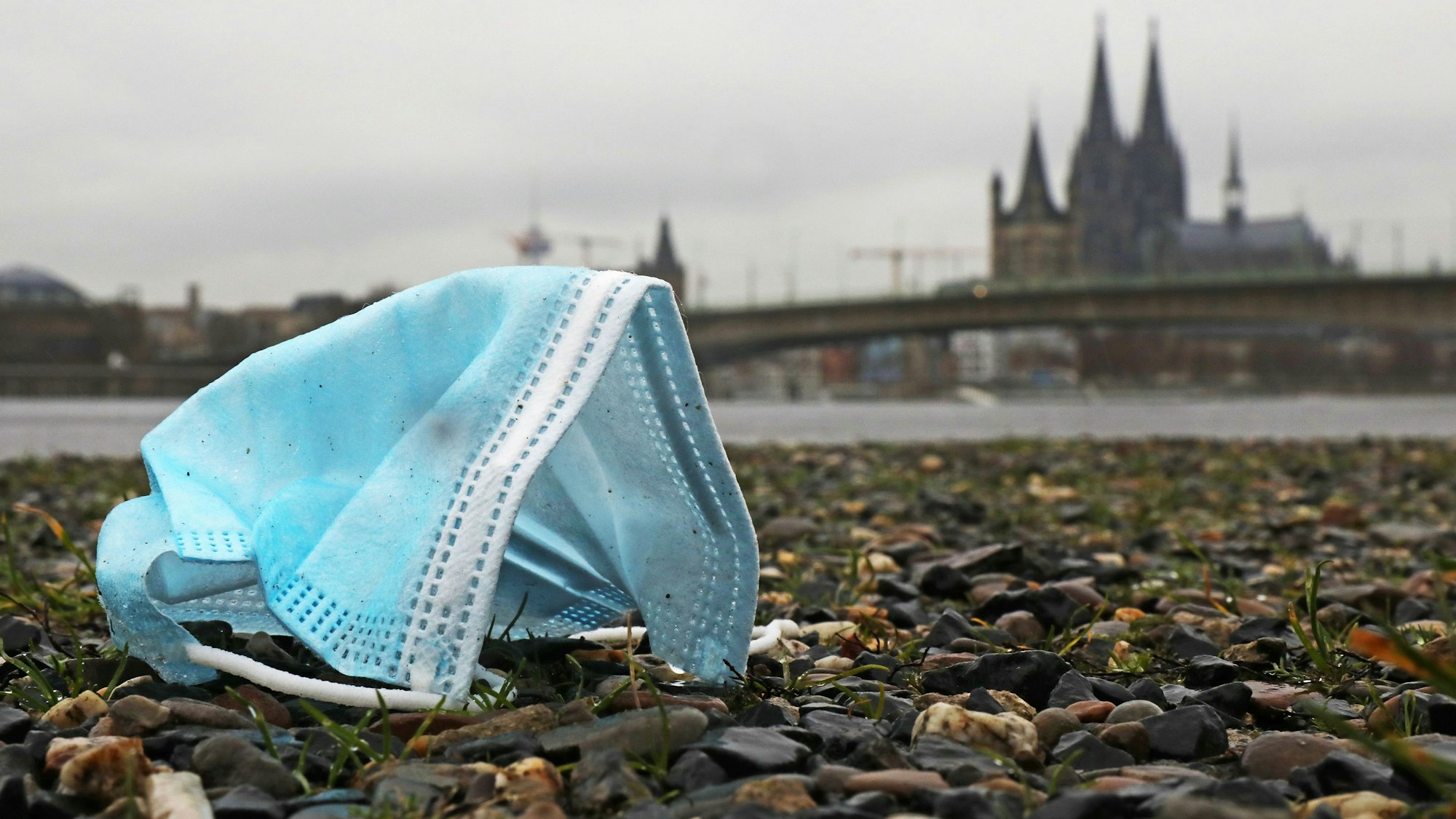 Eine weggeworfene Maske liegt am Rheinufer auf dem Boden. Im Hintergrund sieht man unscharf den Dom.