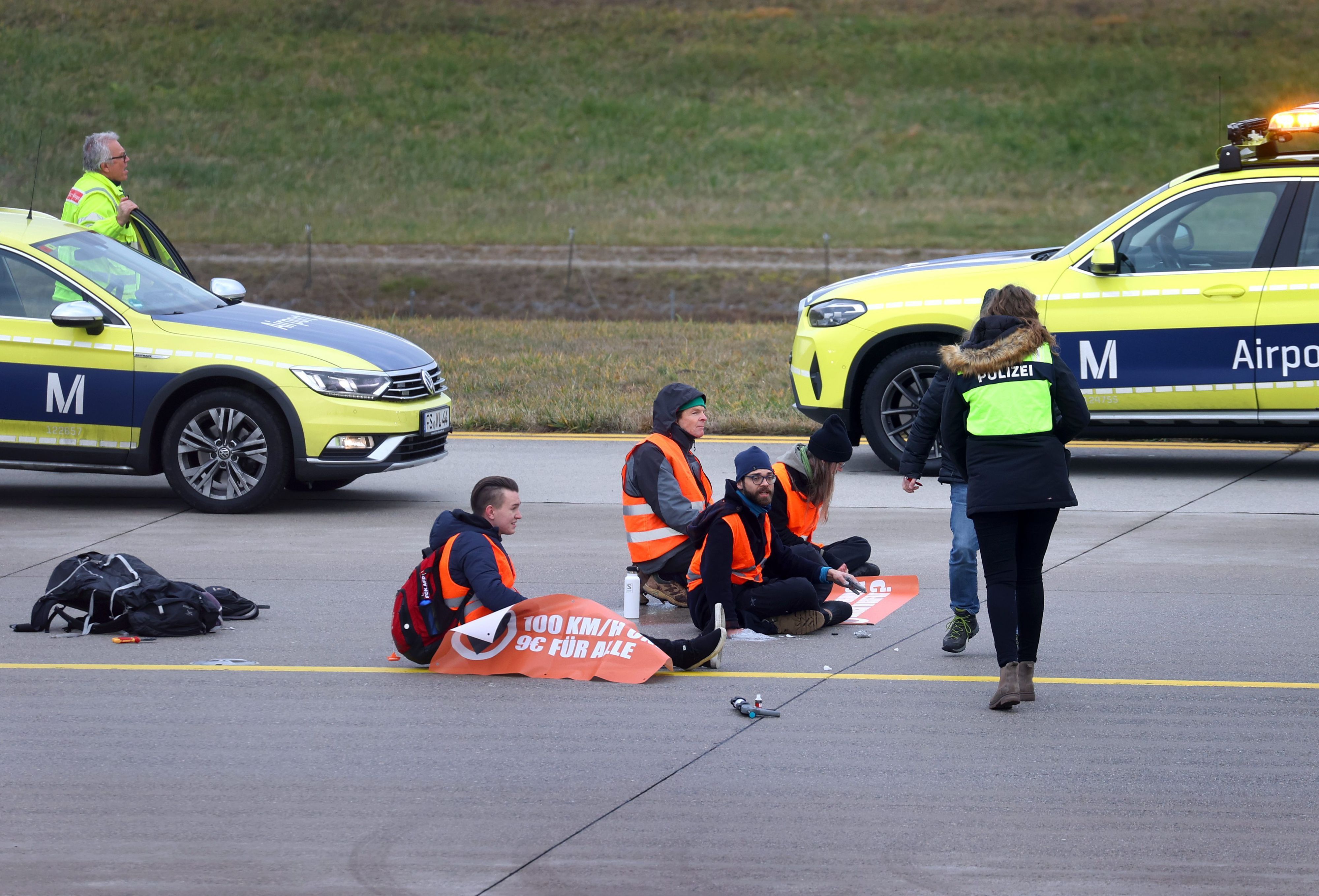 Vier Klimaaktivisten sitzen mit angeklebten Händen auf dem Zubringer einer Start-und Landebahn an einem Flughafen