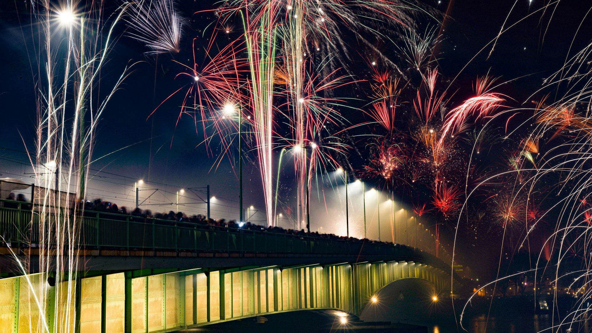 Zahlreiche Menschen stehen am Geländer der Deutzer Brücke während das Feuerwerk den Nachthimmel erleuchtet.