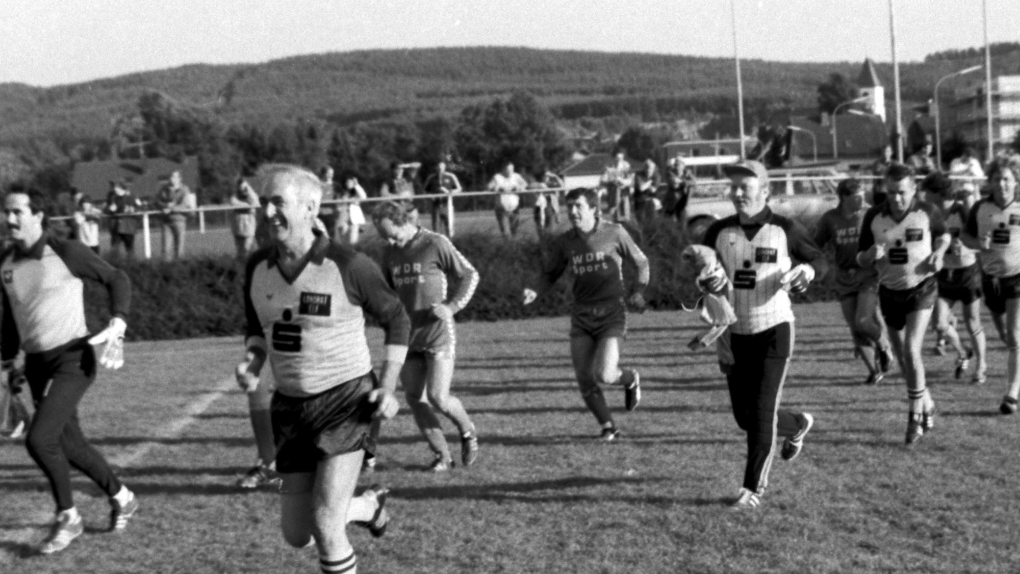 Die Mannschaften des WDR und der Linden-Elf laufen in zwei Reihen nebeneinander im Kaller Grenzlandstadion ein. Die Kreis-Kicker werden von Werner Schumacher (Vordergrund) angeführt.