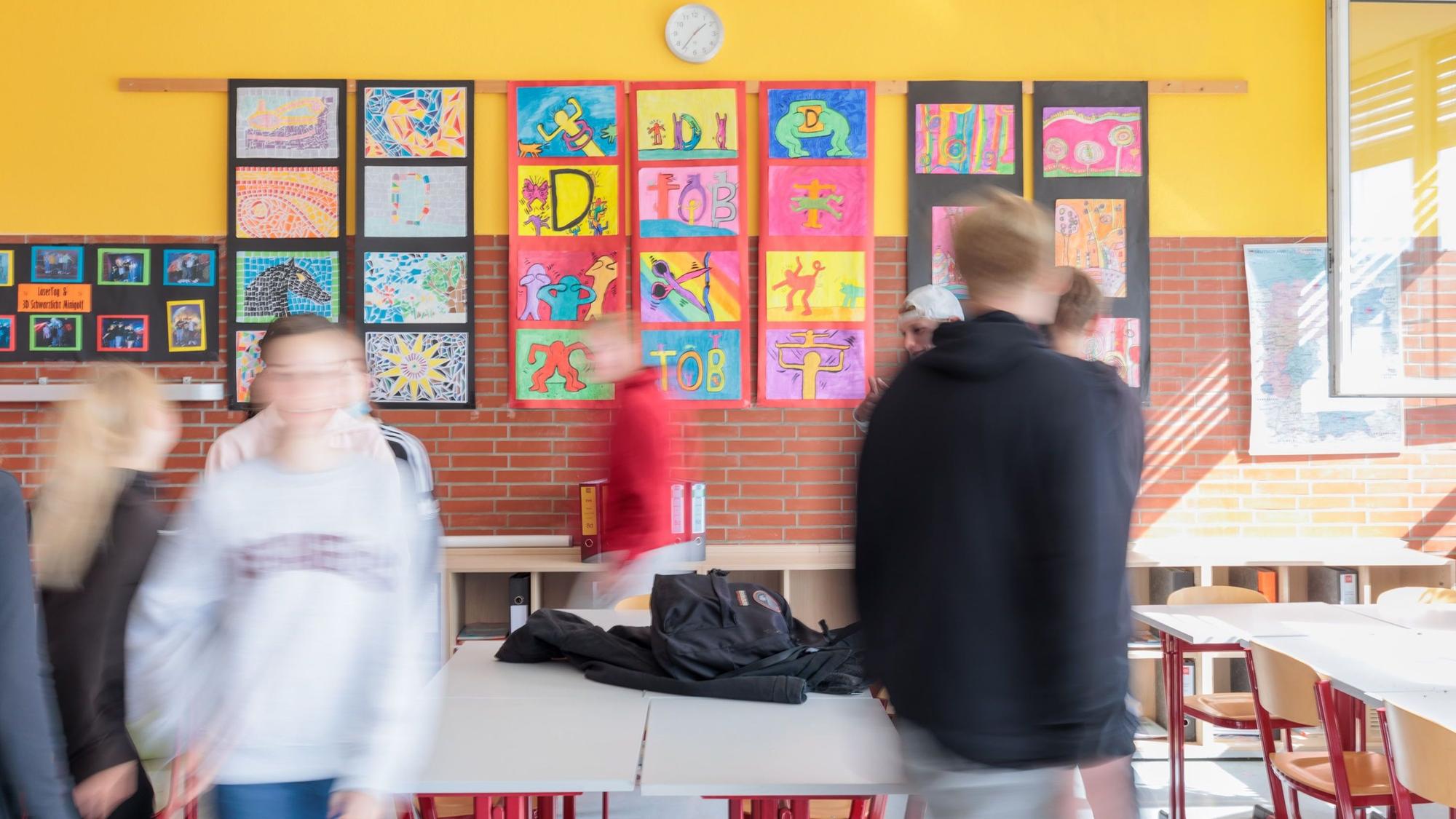 Schülerinnen und Schüler in einem Klassenzimmer mit bunten Bildern an den Wänden. Die Kinder sind verschwommen fotografiert und unkenntlich.