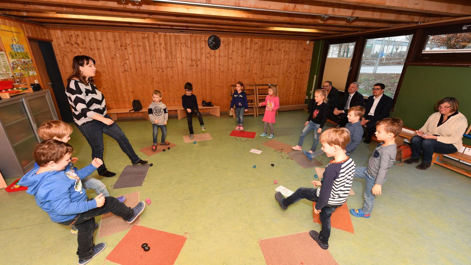Kinder und eine Erzieherin stehen in einem Kreis und heben ihre Beine.