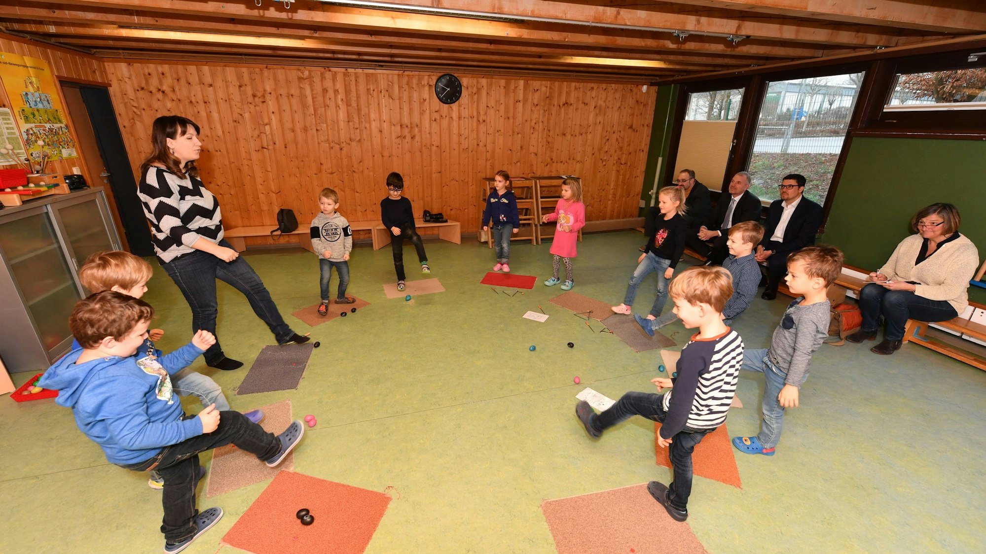 Kinder und eine Erzieherin stehen in einem Kreis und heben ihre Beine.