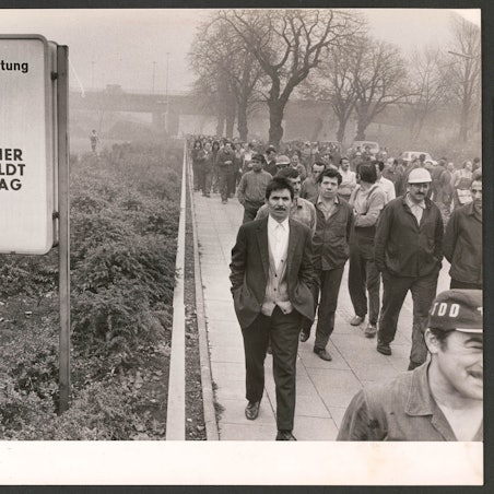 Männer gehen auf dem Schwarz-weiß-Foto in Arbeitskleidung in einem Streikmarsch am Torschild des Werksgeländes vorbei.