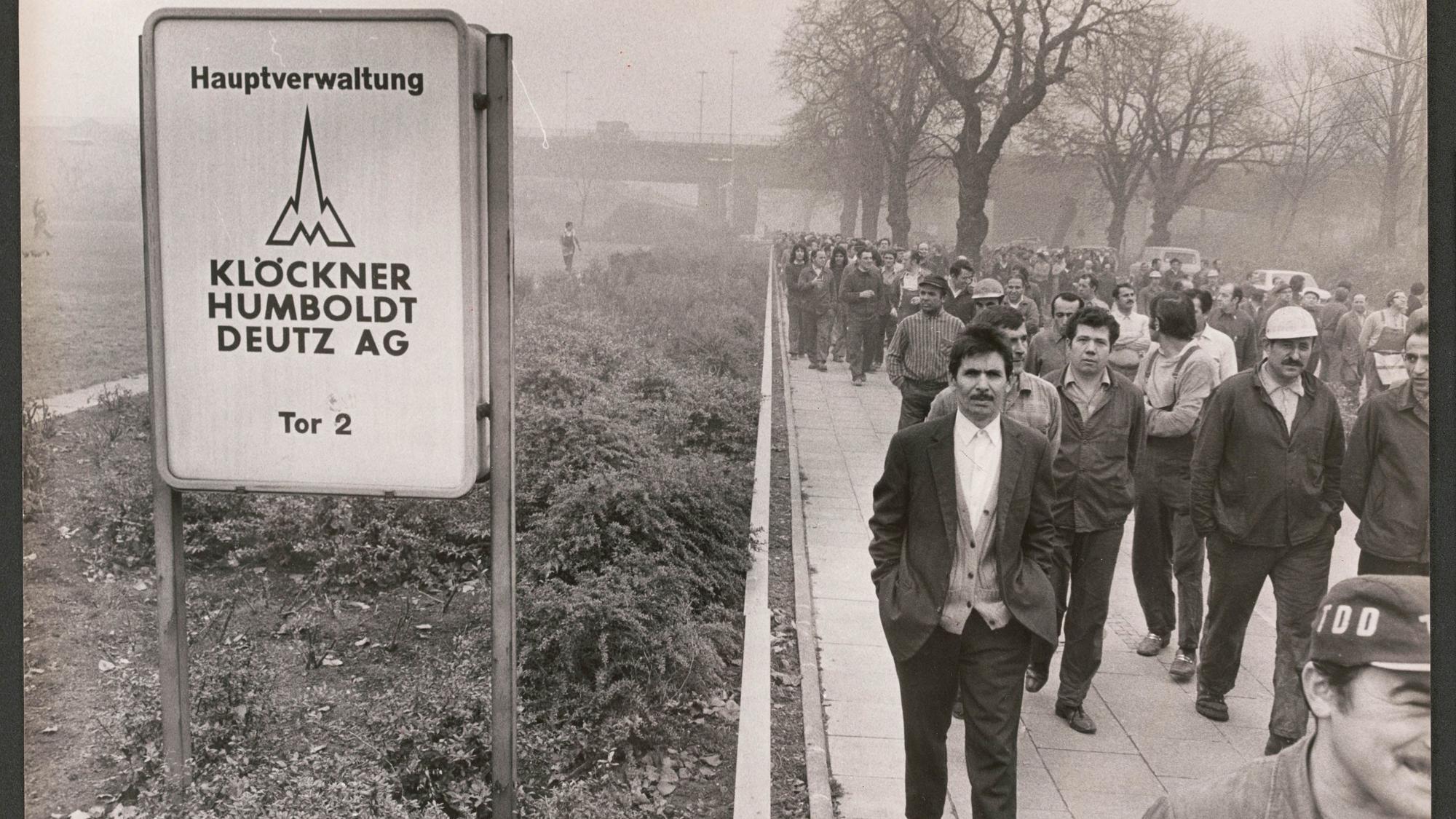Männer gehen auf dem Schwarz-weiß-Foto in Arbeitskleidung in einem Streikmarsch am Torschild des Werksgeländes vorbei.