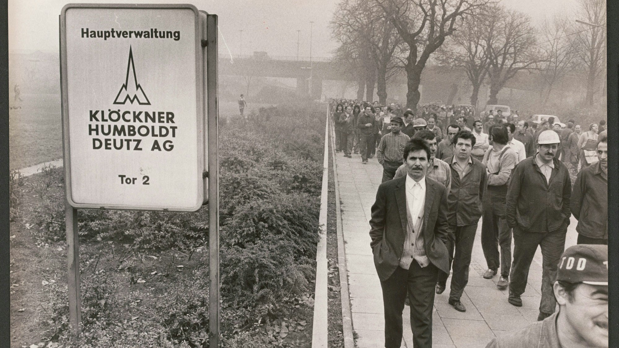 Männer gehen auf dem Schwarz-weiß-Foto in Arbeitskleidung in einem Streikmarsch am Torschild des Werksgeländes vorbei.
