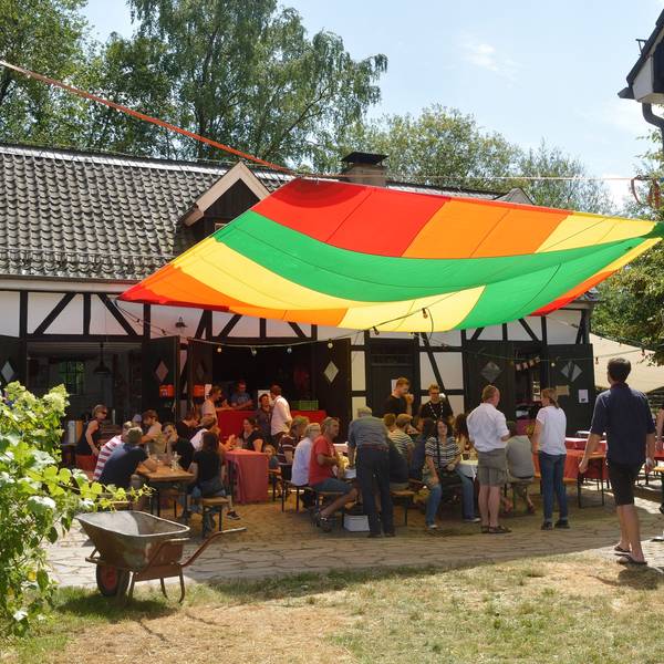 Menschen sitzen vor dem Kochshof und unter einem bunten Segel beim Patentreffen 2019.