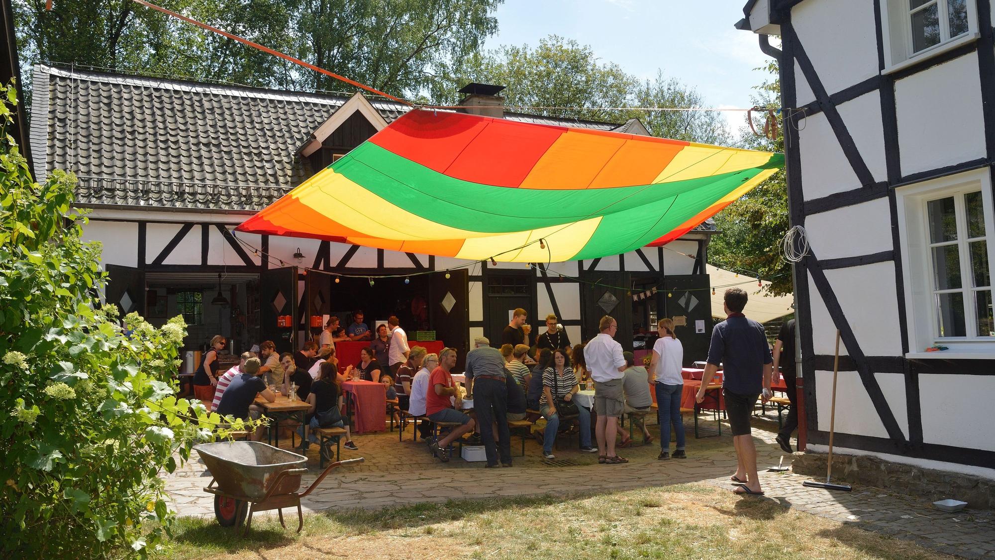 Menschen sitzen vor dem Kochshof und unter einem bunten Segel beim Patentreffen 2019.