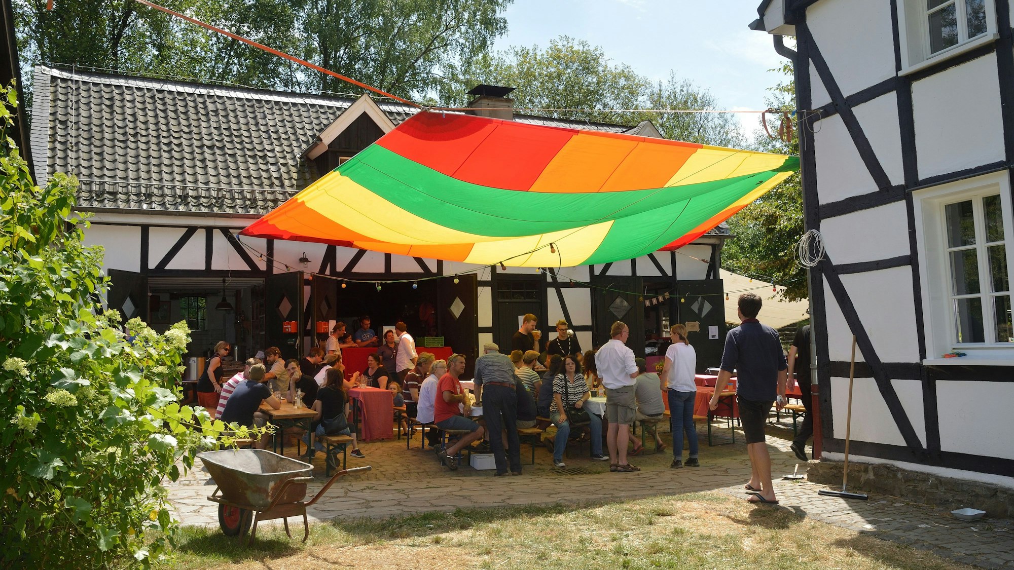 Menschen sitzen vor dem Kochshof und unter einem bunten Segel beim Patentreffen 2019.