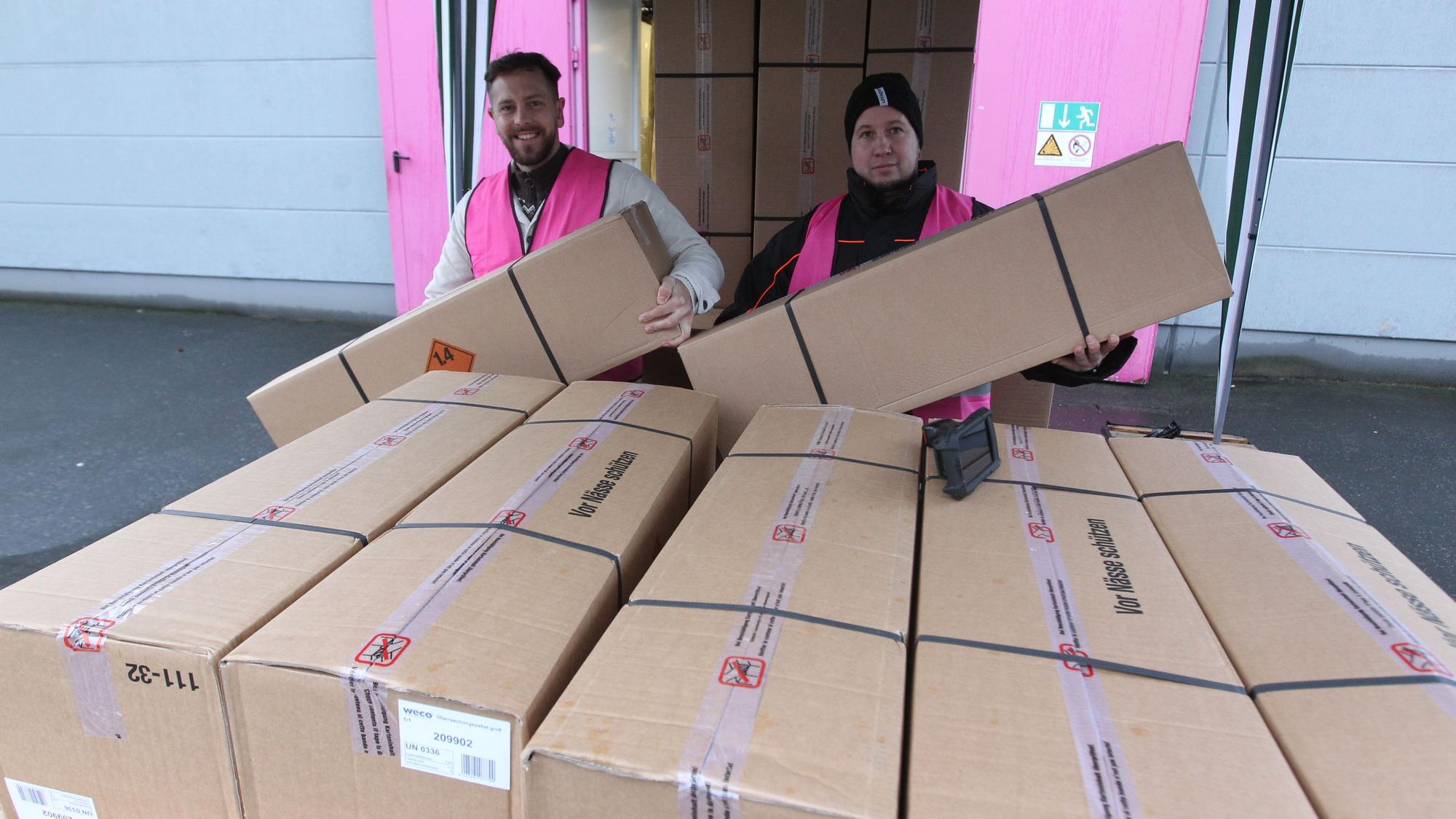 Zwei Männer in pinkfarbenen Weco-Westen heben zwei lange Pappkartons in die Höhe, vor ihnen steht ein hoher Stapel weiterer Kartons.