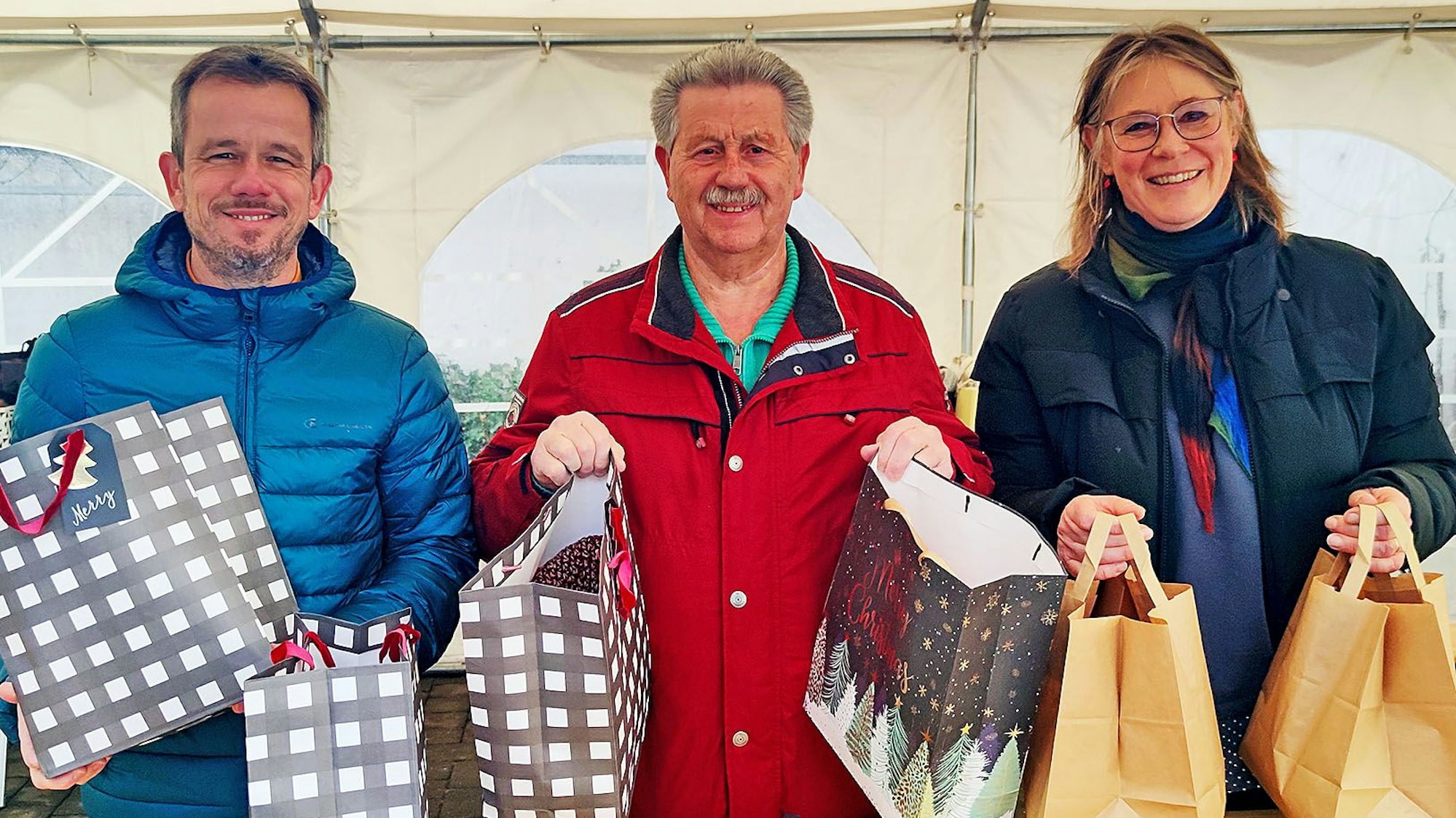 Martin Jost, Hubert Hamacher und Maria Surges-Brilon stehen unter einem Zelt und halten Weihnachtstüten in den Händen.