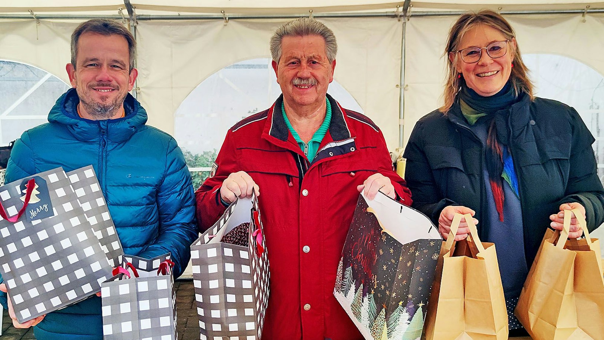 Martin Jost, Hubert Hamacher und Maria Surges-Brilon stehen unter einem Zelt und halten Weihnachtstüten in den Händen.