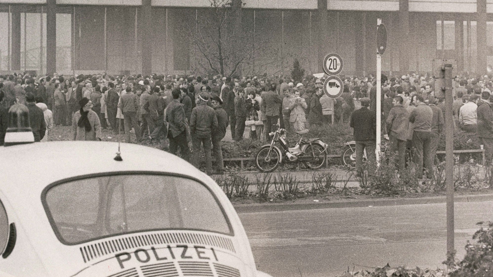 Streikende vor einem KHD-Gebäude unter Beobachtung durch die Polizei. Im Vordergrund des Schwarz-weiß-Fotos ist ein weißer VW-Käfer mit Polizeiaufschrift zu sehen.