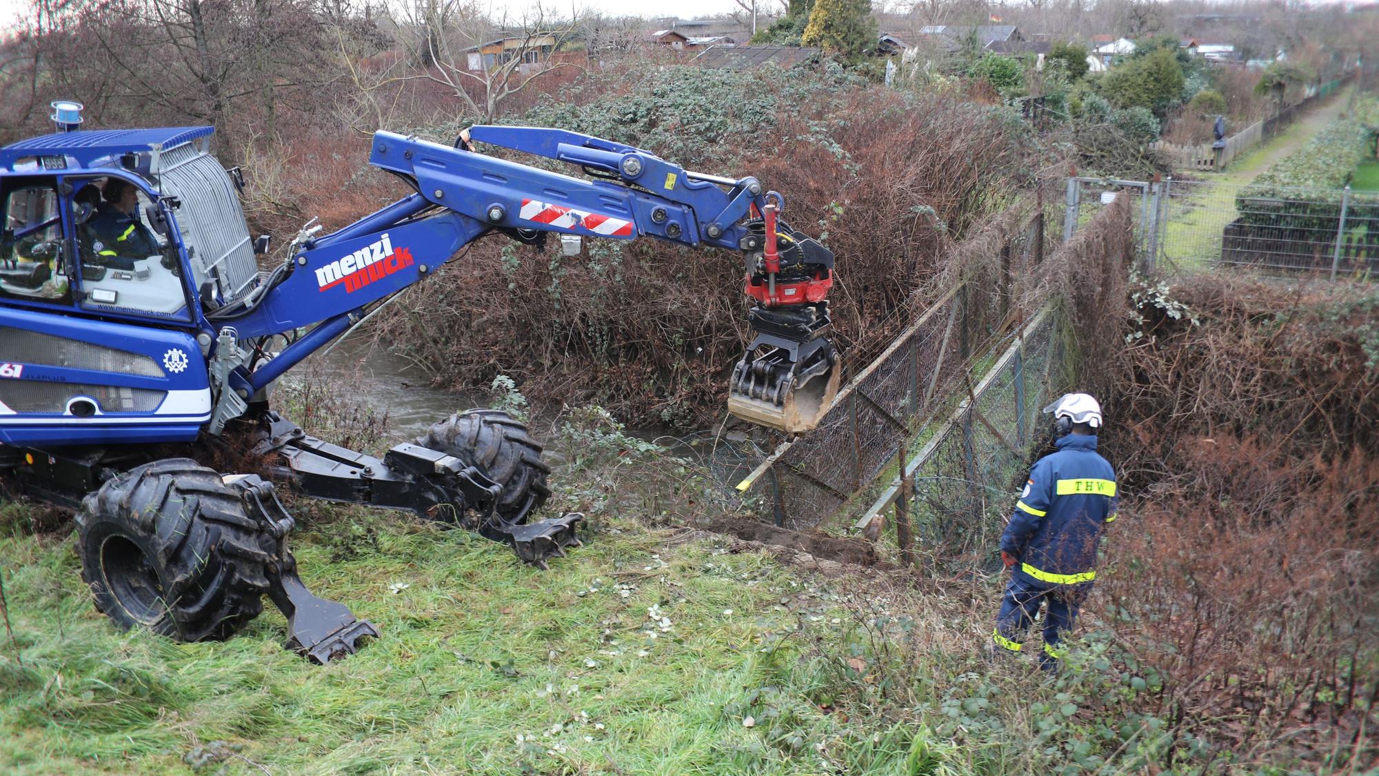 Ein blauer Bagger steht vor einer kleinen Brücke über einen Graben, die abgerissen wird.
