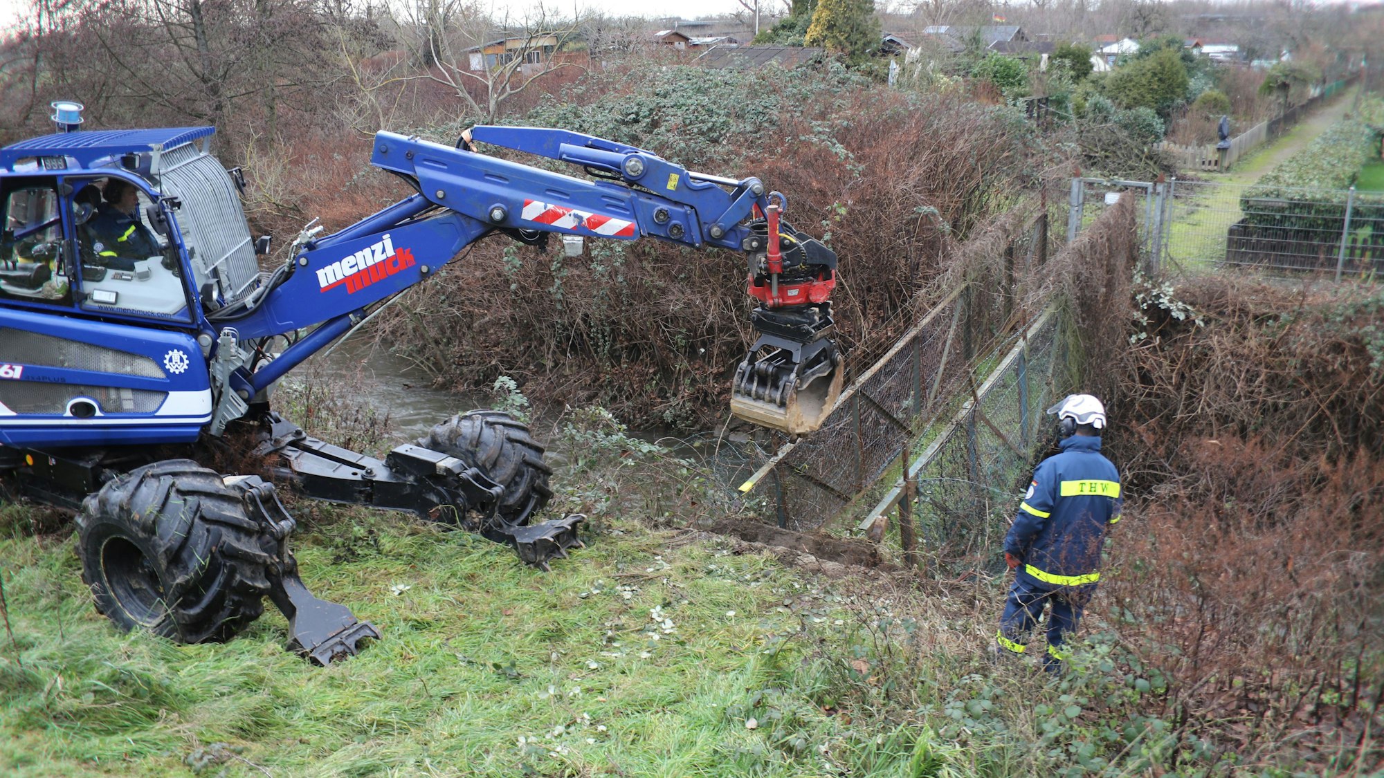 Ein blauer Bagger steht vor einer kleinen Brücke über einen Graben, die abgerissen wird.