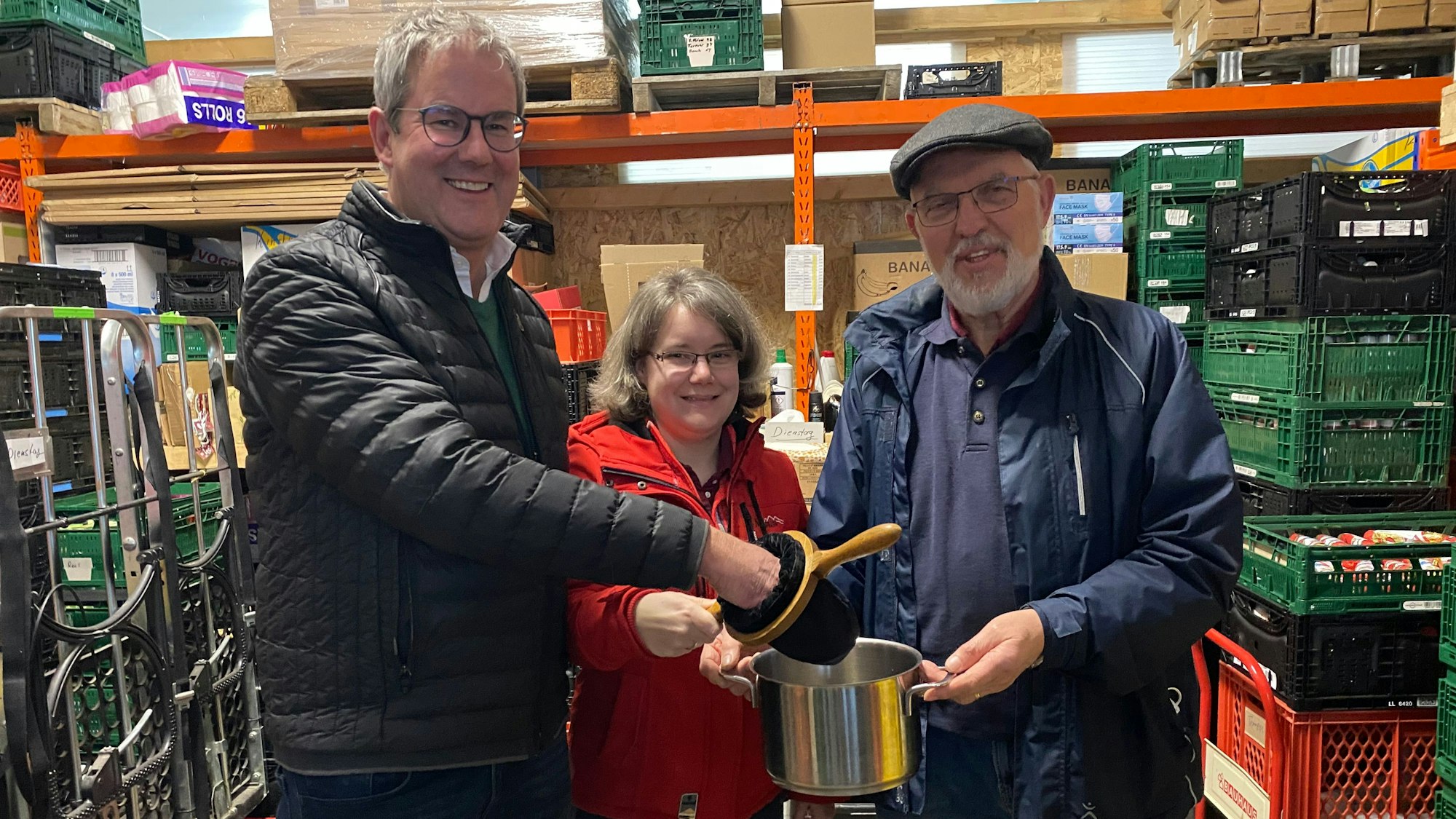 Pfarrer Frank Thönnes (v.l.), Tammy Peiter und Walter Feckinghaus stehen im Lager der Tafel Euskirchen und halten den Klingelbeutel in der Hand, der symbolisch in einem Topf entleert wird.