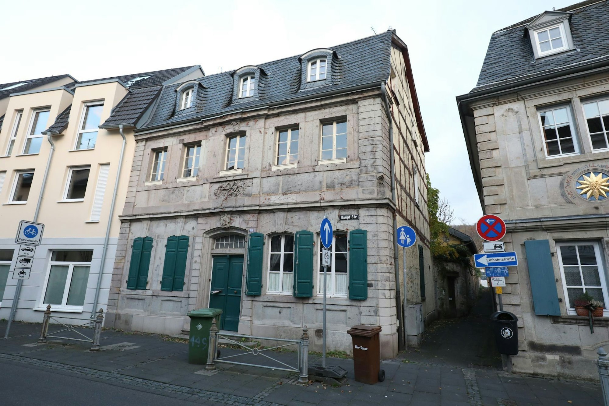 Das Bild zeigt einen barocken Altbau mit grauer Werksteinfassade, grüner Tür und grünen Fensterläden in der Hauptstraße in Königswinter; rechts neben dem Gebäude gibt es eine kleine Gasse.