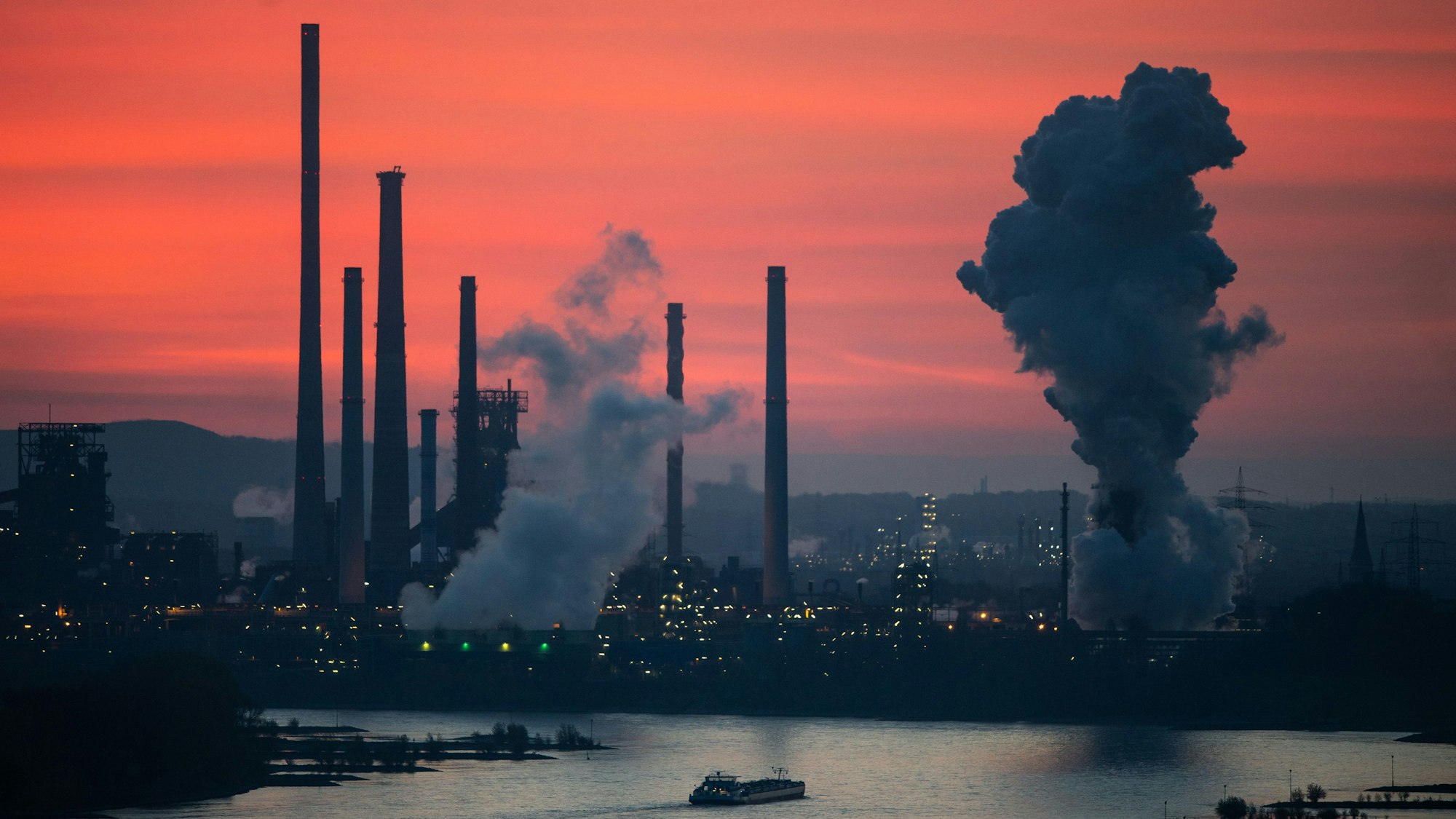 Die Silhouette einer Industrieanlage vor einem rosa Himmel bei Sonnenaufgang. Dampf steigt aus Schornsteinen.
