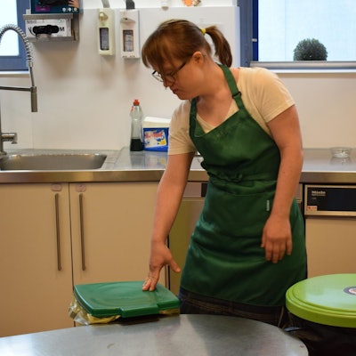 Anna Krechel steht in grüner Schürze in einer Küche neben den Mülleimern.