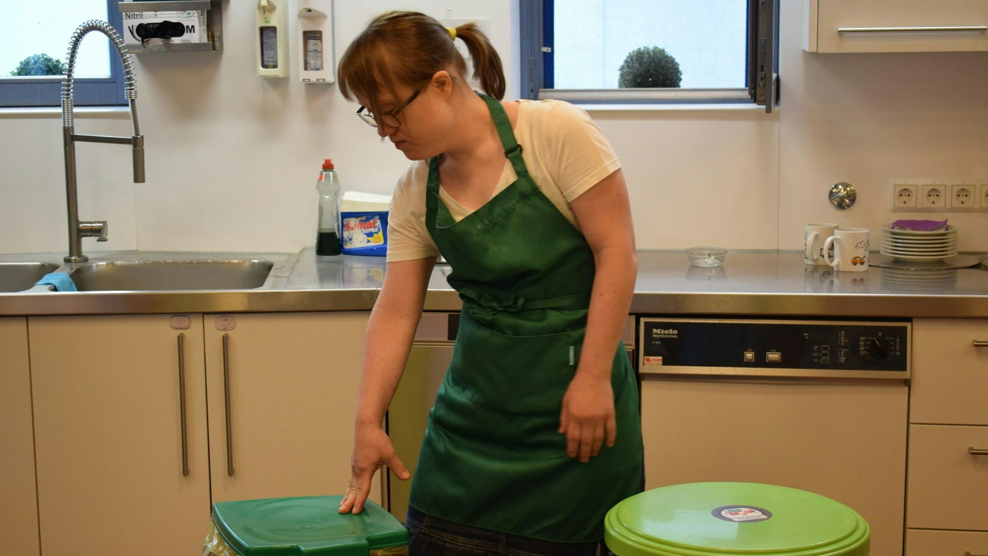 Anna Krechel steht in grüner Schürze in einer Küche neben den Mülleimern.