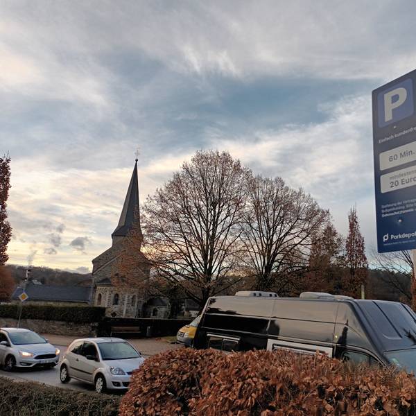 Ein kleines, blaues Schild auf einem Supermarkt-Parkplatz im Lohmarer Zentrum informiert über die Höchstdauer fürs Parken und das Verwarngeld von mindestens 20 Euro. Im Hintergrund ist die katholische Kirche zu sehen.