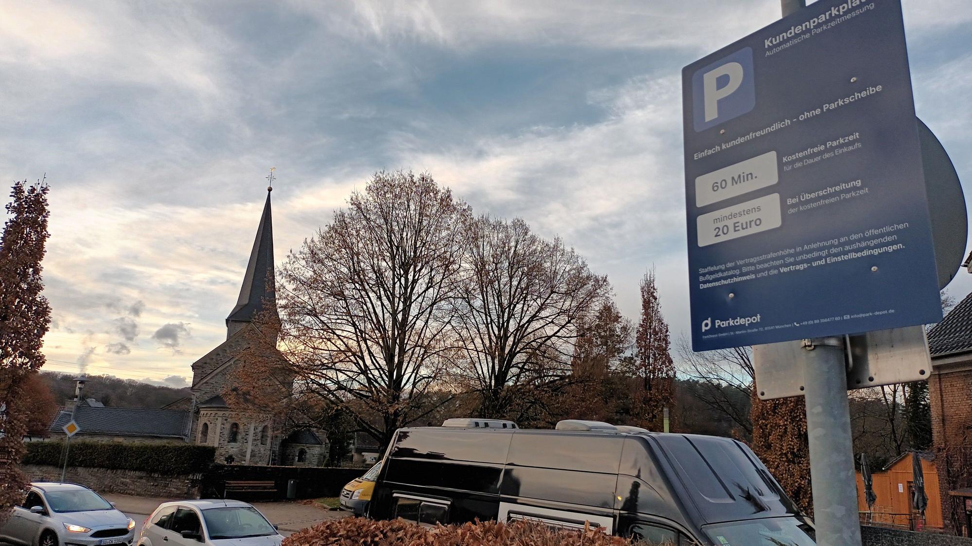 Ein kleines, blaues Schild auf einem Supermarkt-Parkplatz im Lohmarer Zentrum informiert über die Höchstdauer fürs Parken und das Verwarngeld von mindestens 20 Euro. Im Hintergrund ist die katholische Kirche zu sehen.