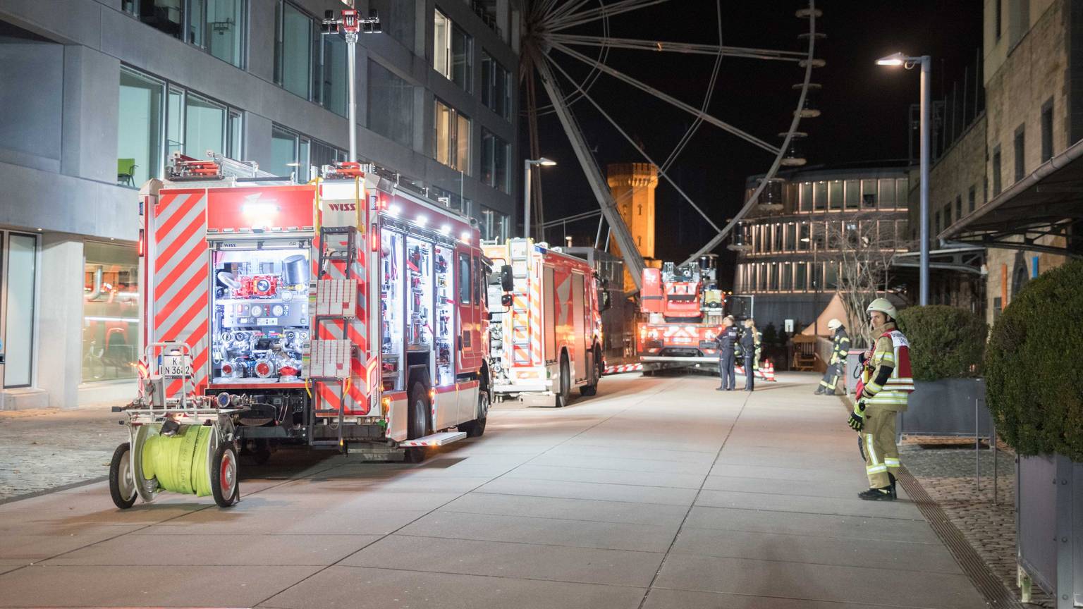 Die Kölner Feuerwehr sichert einen Brand in der Straße "Am Zollhafen" im Kölner Rheinauhafen. Im Hintergrund ist das Riesenrad am Schokoladenmuseum zu sehen.