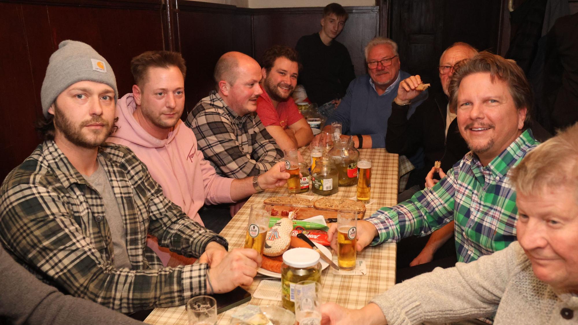 Eine Männerrunde sitzt an einem langen Tisch und spielt Dobbeln. Auf dem Tisch stehen Biergläser und Teller, auf denen Wüste liegen.